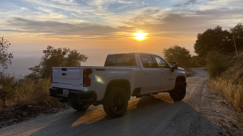 Rear 3/4 view of a white Chevrolet Silverado 2500 HD Trail Boss parked on a dirt road in front of a sunset