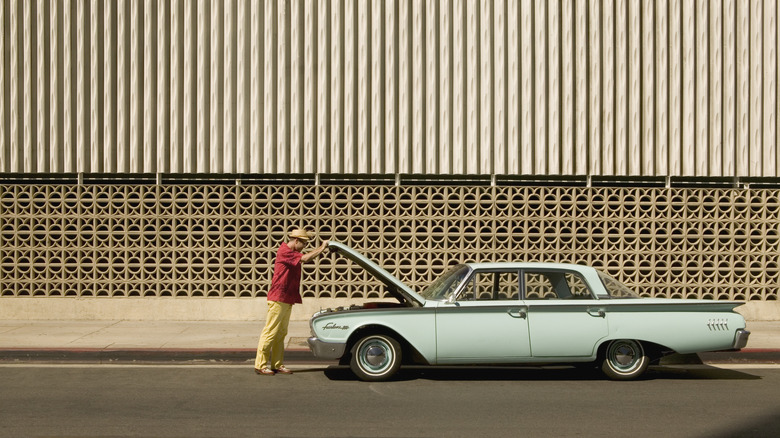 A guy in a red shirt and khaki pants looking under the hood of his classic car.