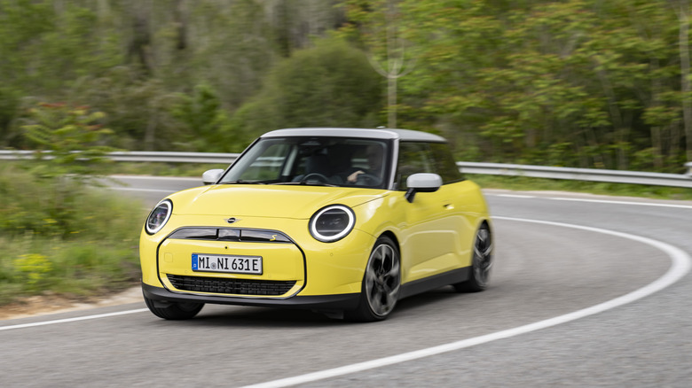 A yellow Mini Cooper SE driving around a turn in front of trees