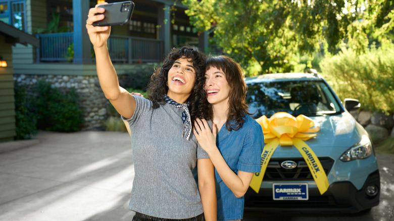 A couple taking a selfie in front of a subaru Crosstrek with a carmax bow on the hood