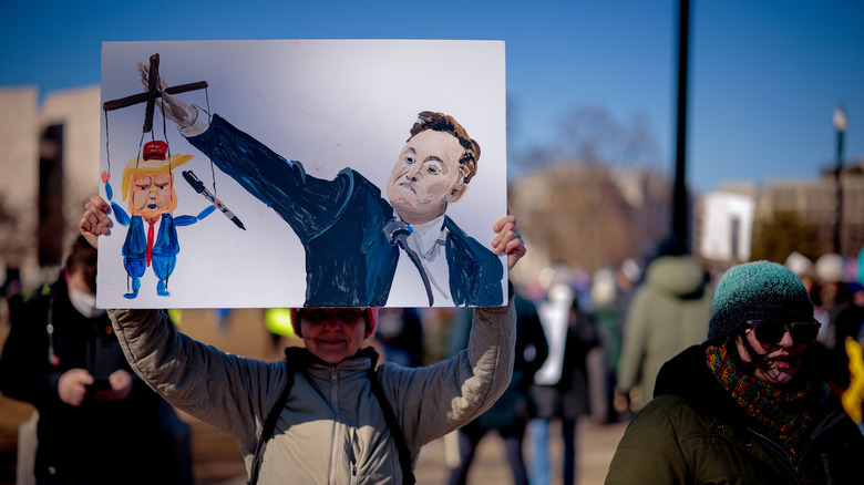 Anti-Elon Musk protestor holds up a sign showing Musk using a right-arm salute to hold a marionette of Trump