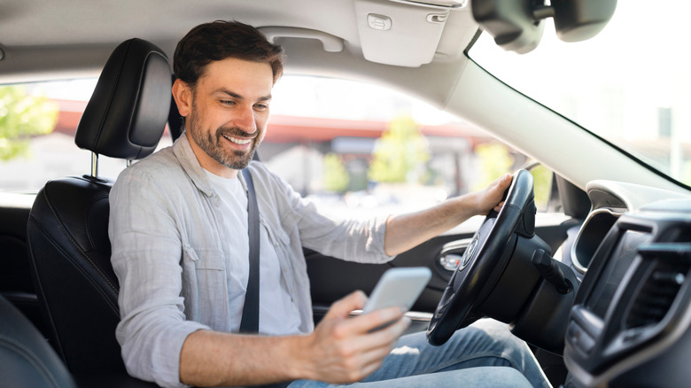 Happy handsome millennial guy driver using smartphone while driving car, using navigation system gps. Businessman chatting with friends or clients on mobile phone while going to office by auto