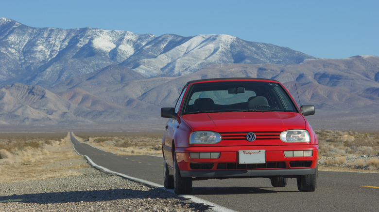 A red 1998 VW Cabrio chugging along a desert highway with mountains in the background