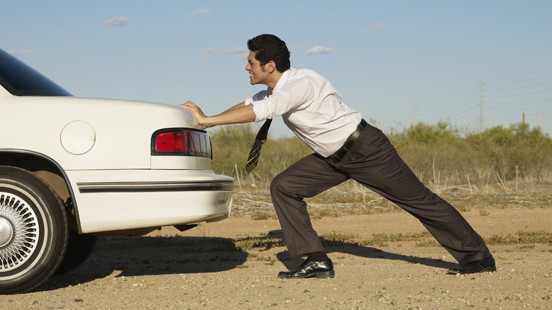 A guy in business clothes pushing a white car on a dirt road.