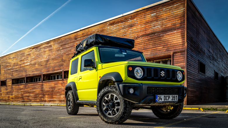 A bright yellow Suzuki Jimny parked next to a wooden building under a clear blue sky.