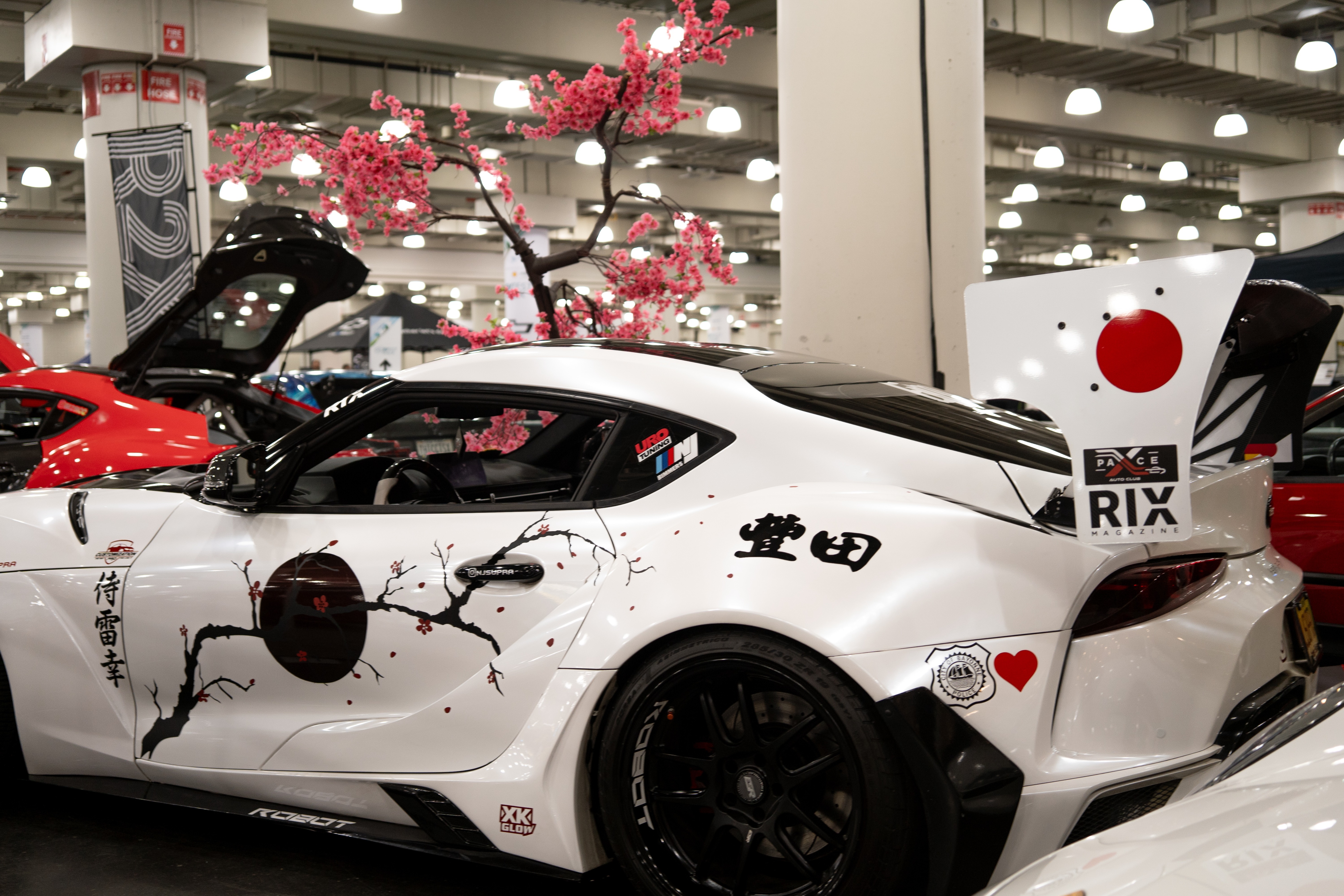 A white Toyota Supra in sakura/Japanese flag livery against a sakura tree