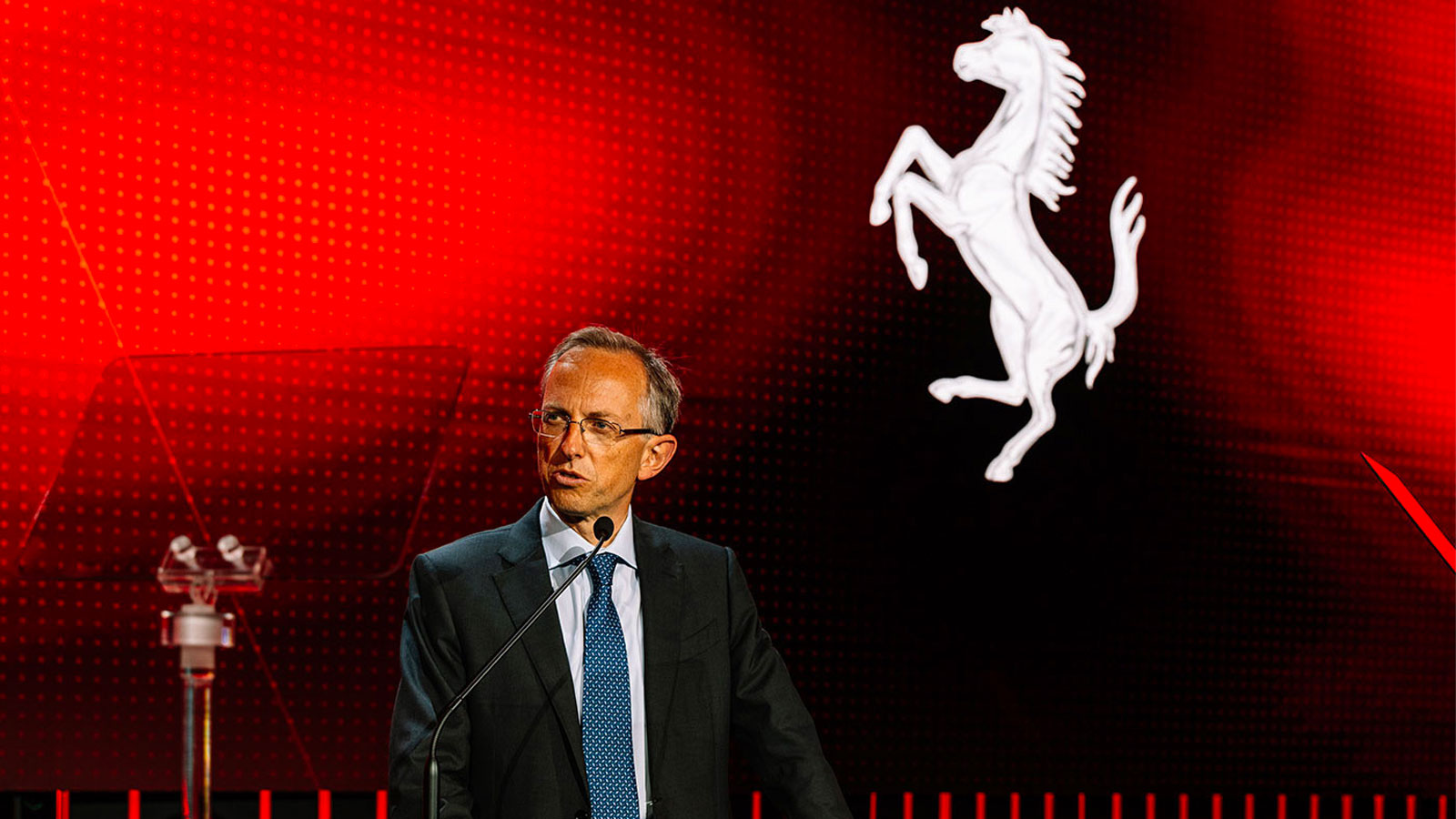 A photo of Ferrari Chief Executive Benedetto Vigna on a red stage with a prancing horse logo in the background. 