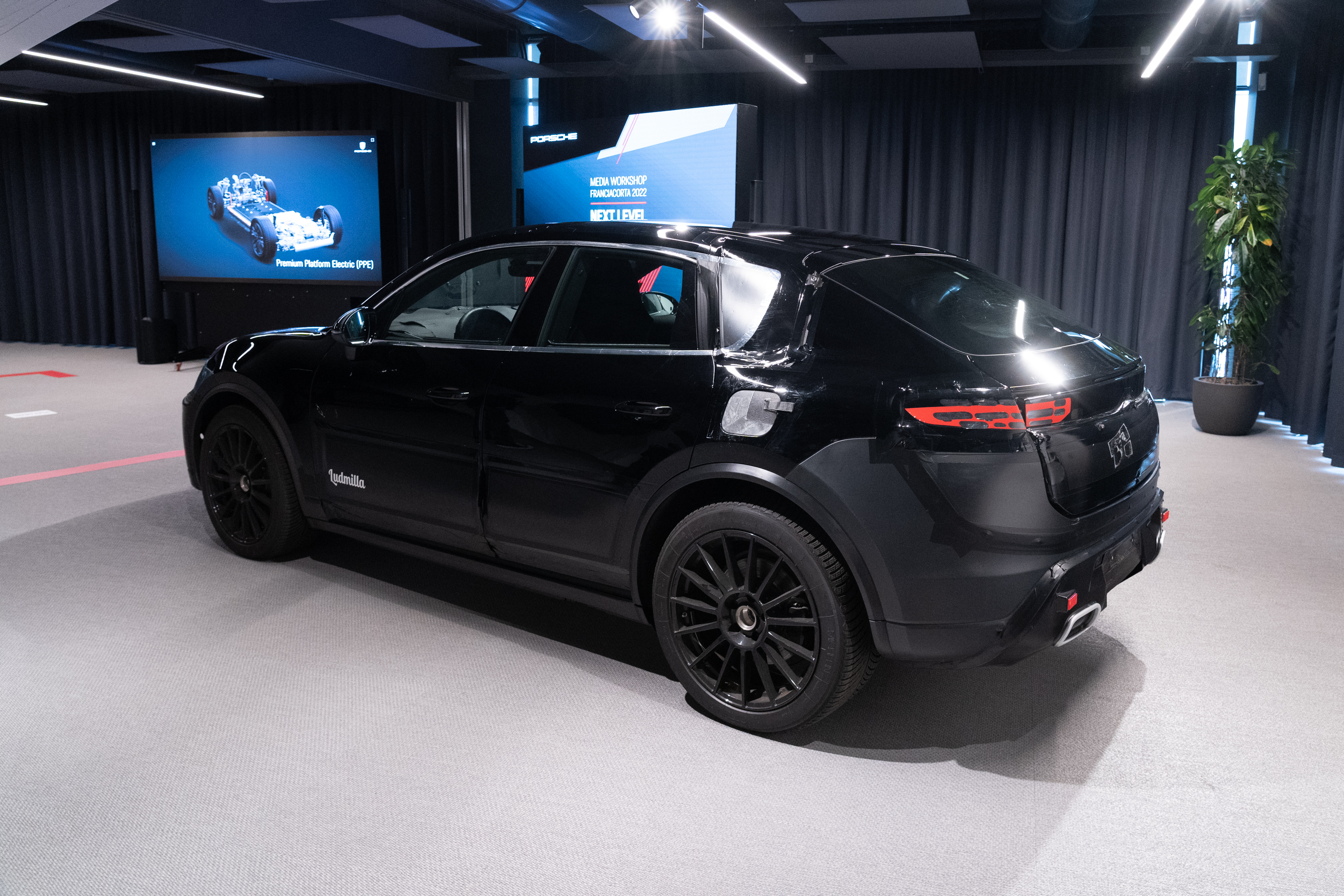 a prototype of the 2024 porsche macan electric photographed inside a convention center. we see the vehicle from the left-rear quarter view. it's painted black with vinyl wrapping to obscure some design details. 