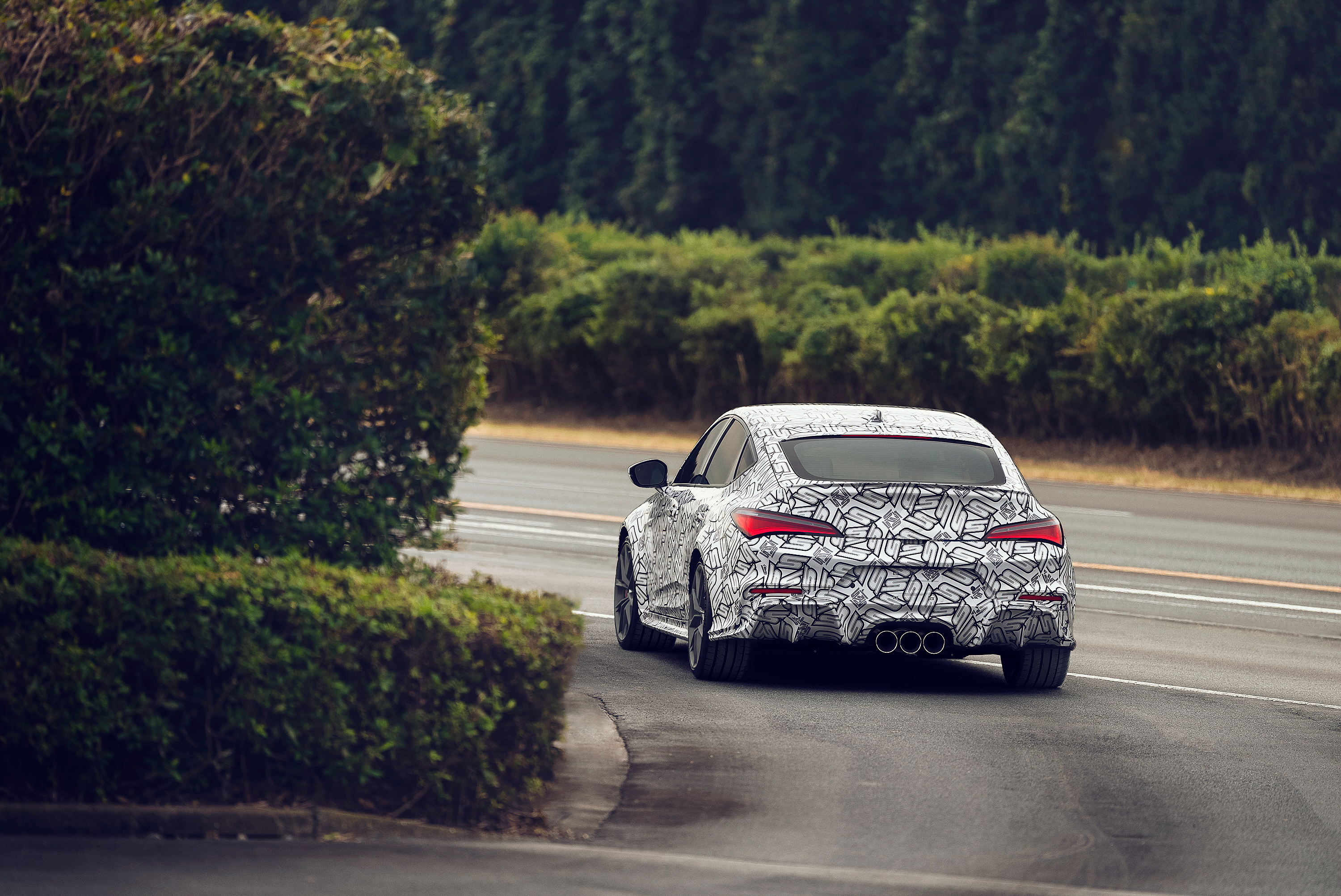 rear view of a 2024 acura integra type s prototype driving away from the camera, going around a left-hand bend on a road flanked by tall bushes on both sides