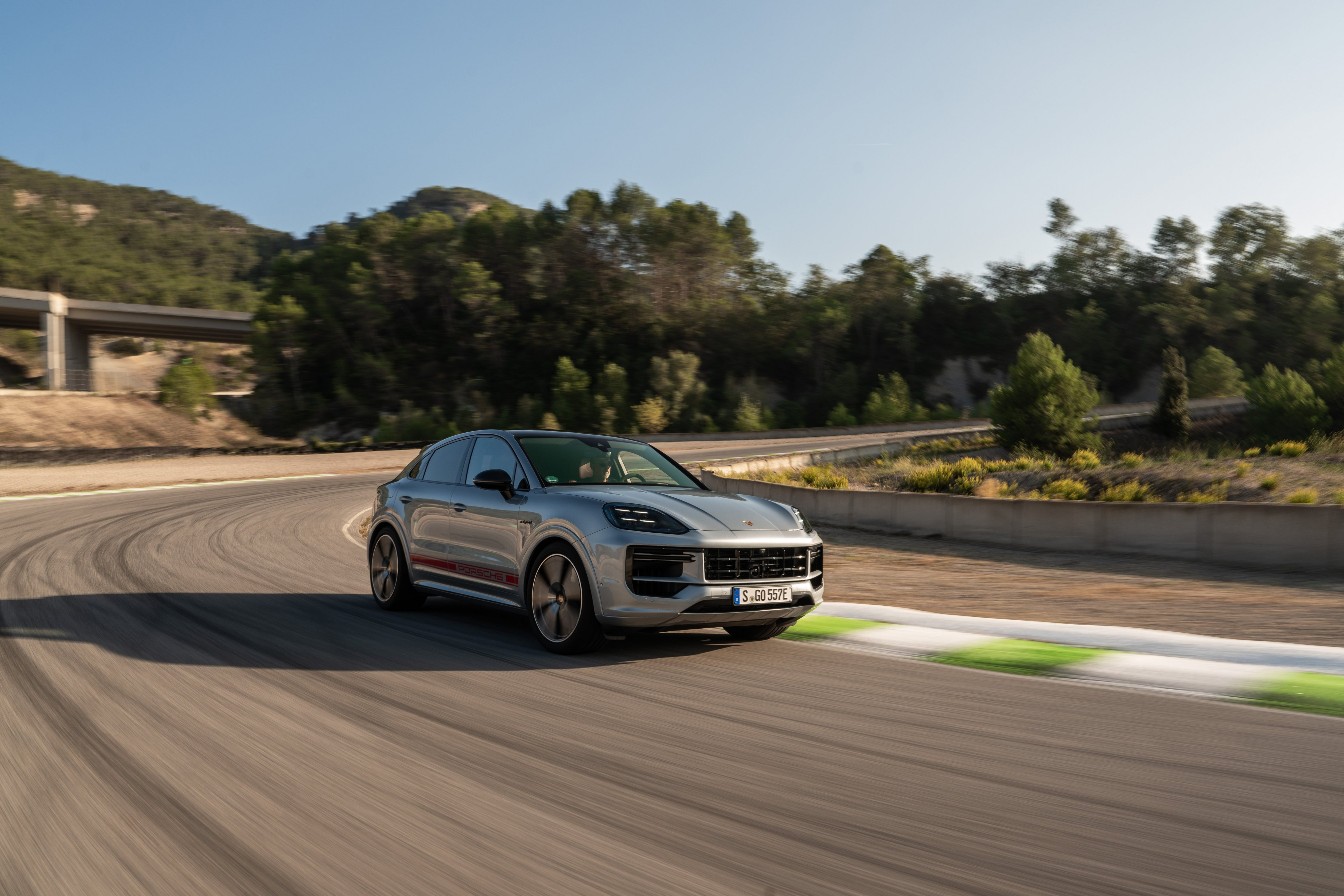 A silver 2024 Porsche Cayenne Turbo E-Hybrid coupe drives swiftly on a track