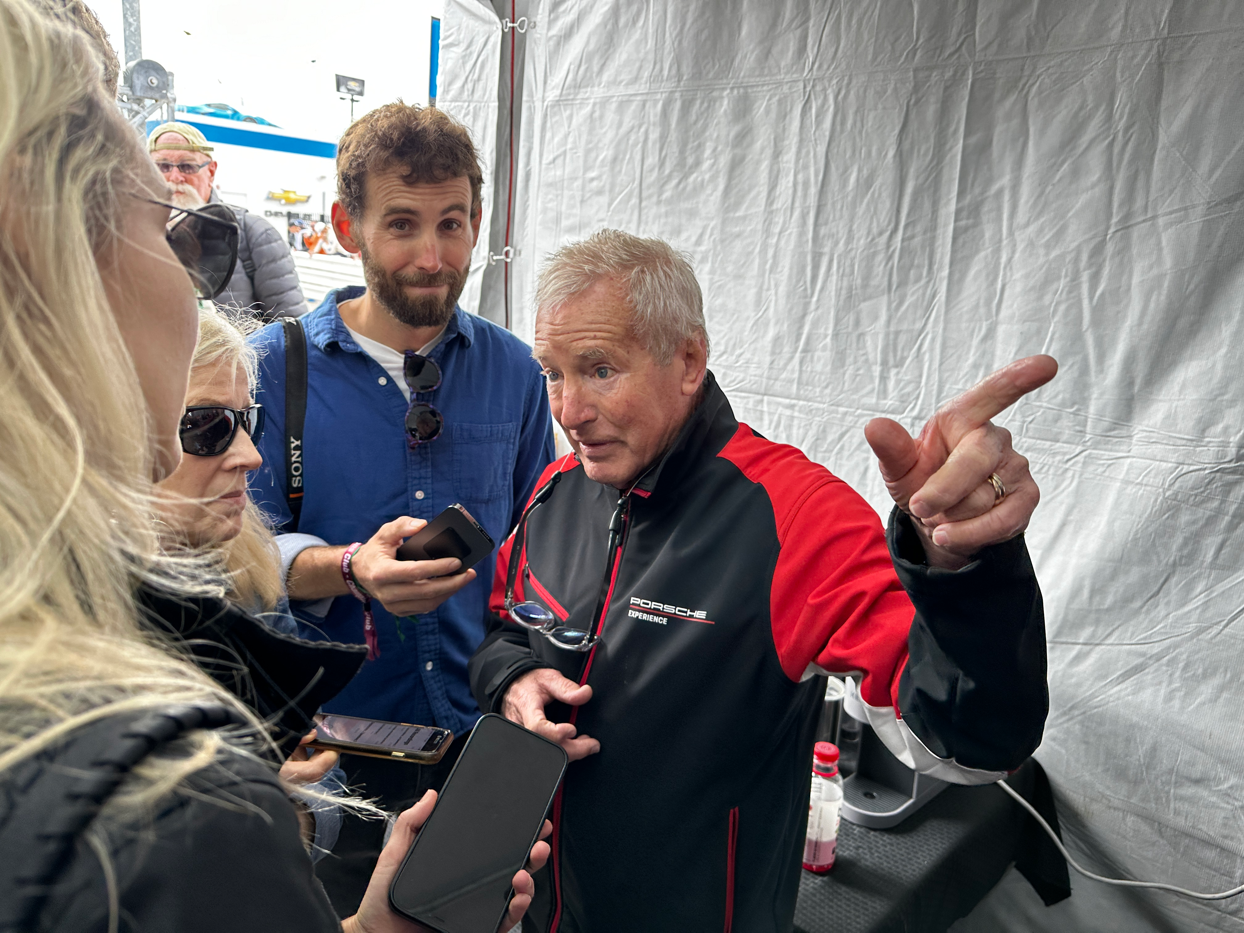 Porsche driver Hurley Haywood points out of the frame while wearing a red and black jacket.