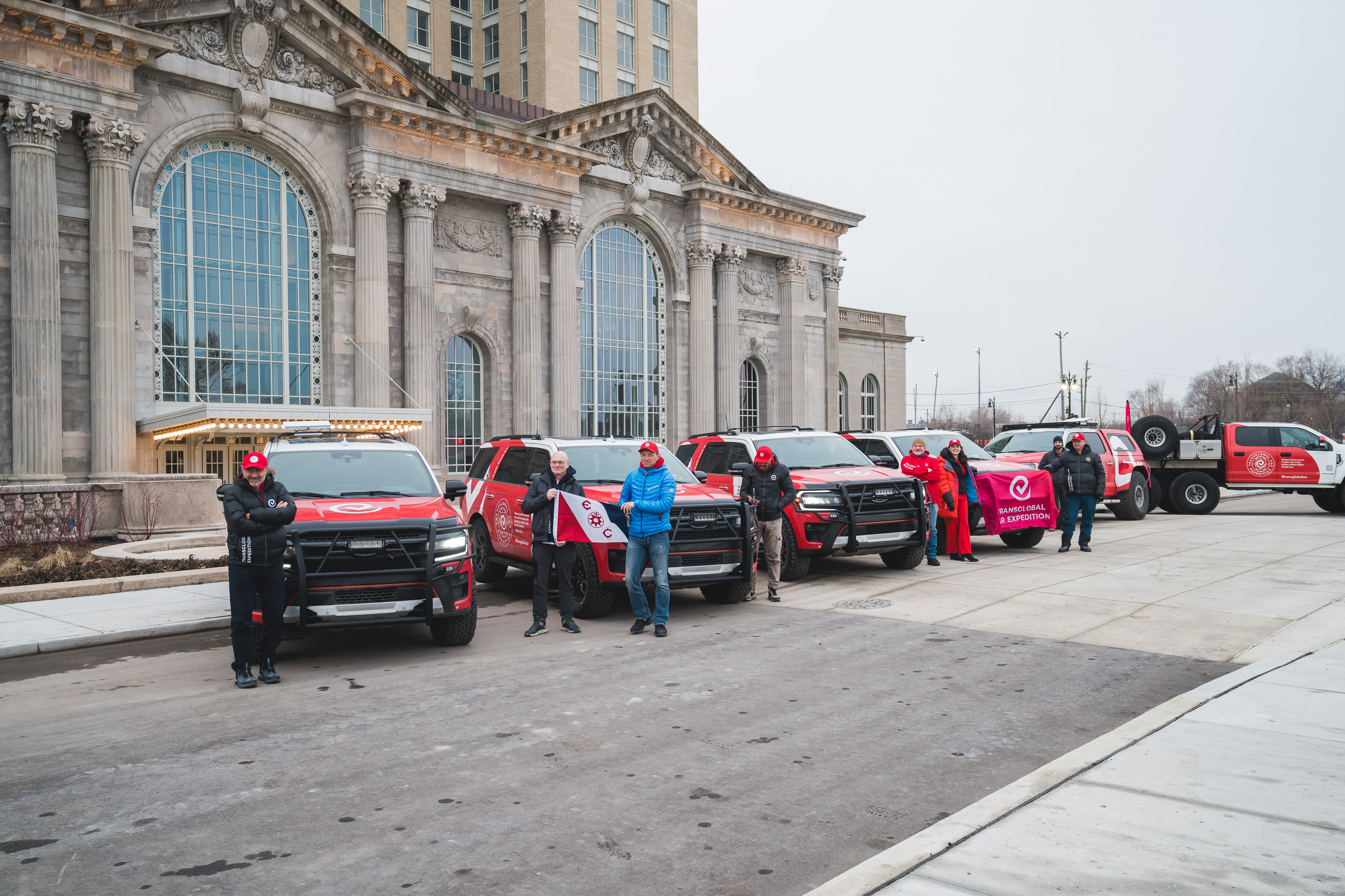 Five red and white F-150s line up in front of an old train station and pose for pictures, with several people standing outside the cars in winter gear holding flags.