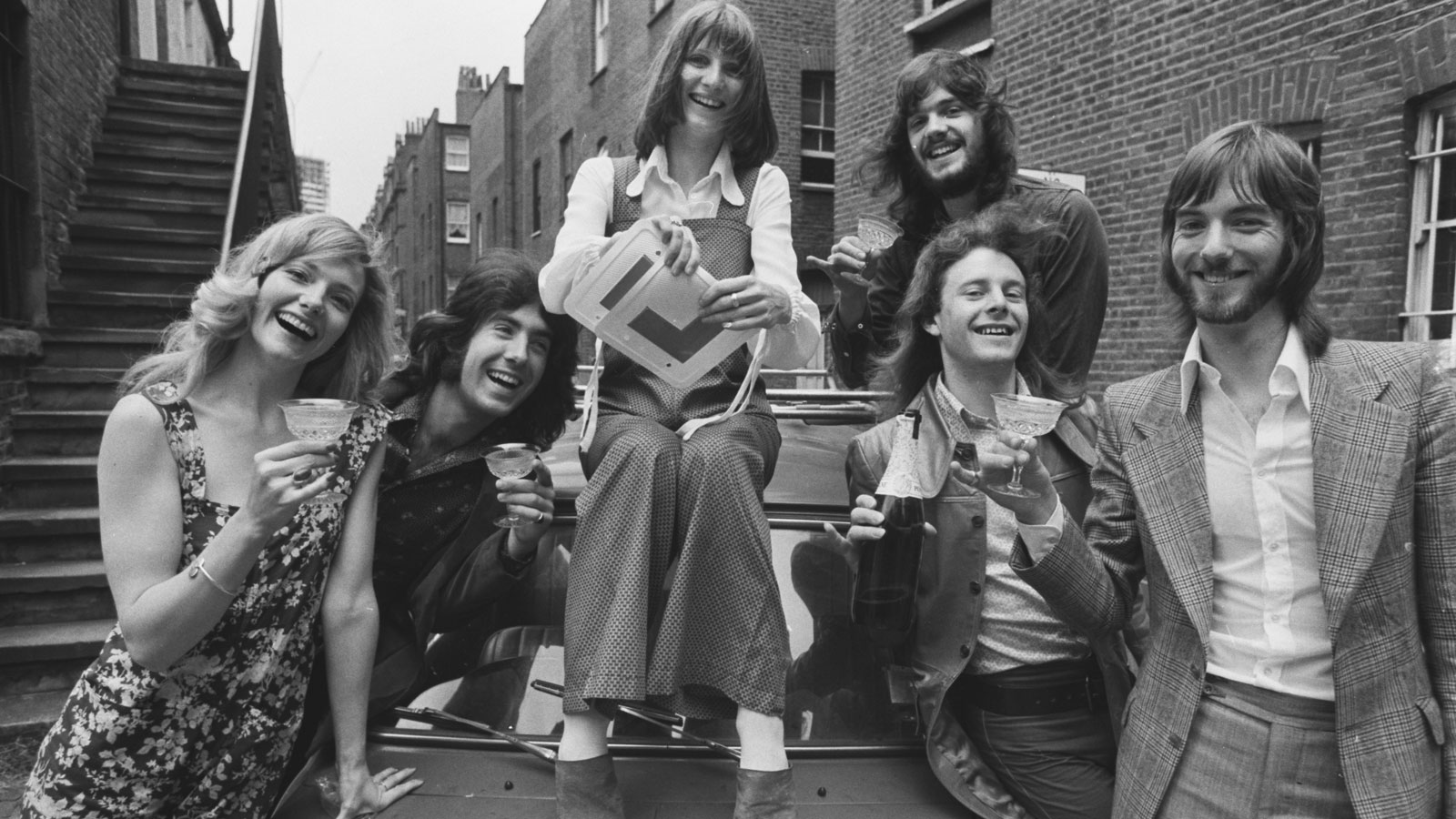 A group of people gather round a car and tear up their L plates. 