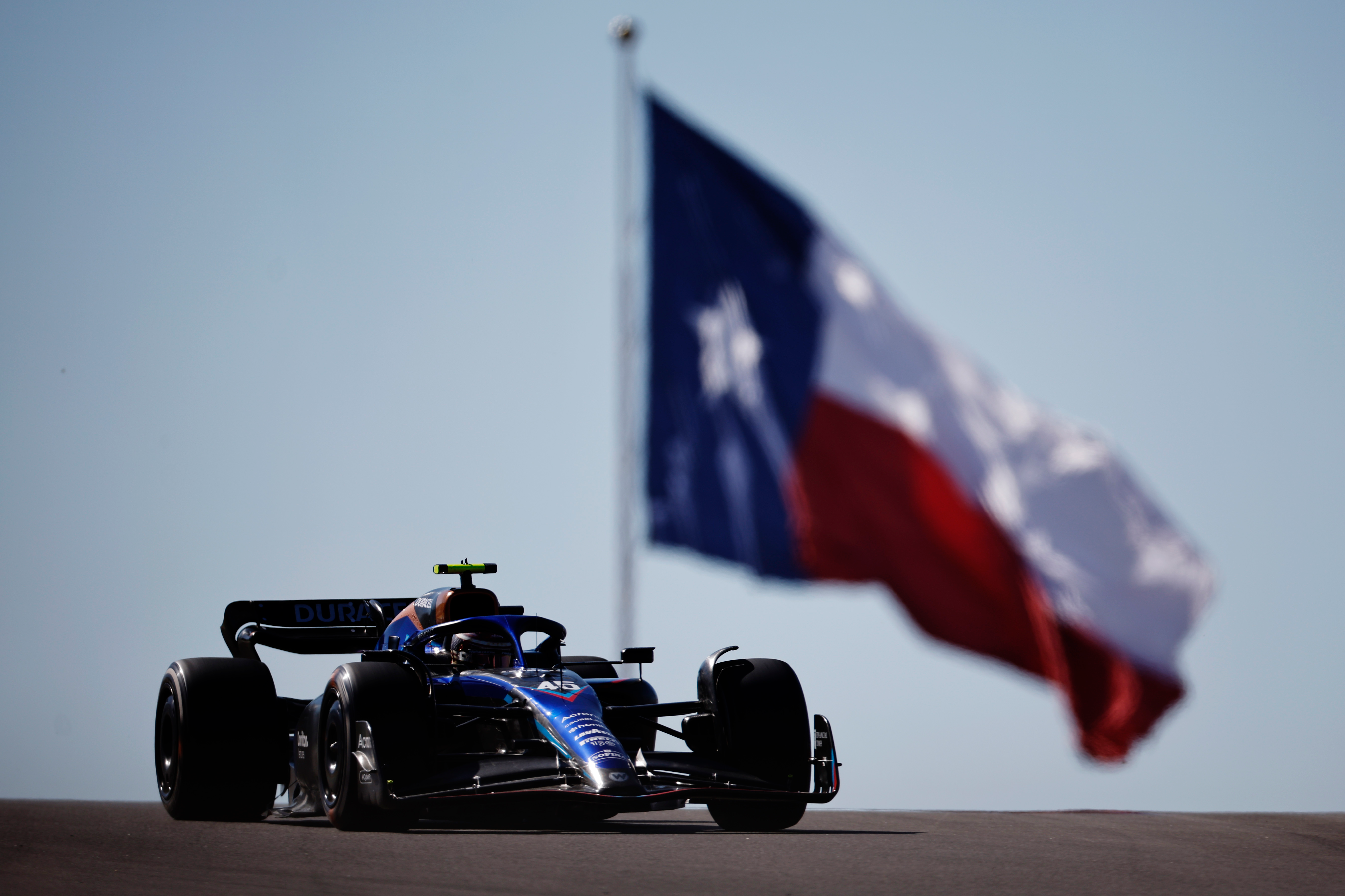 A Williams F1 car driving over a crest with a massive Texas state flag in the background