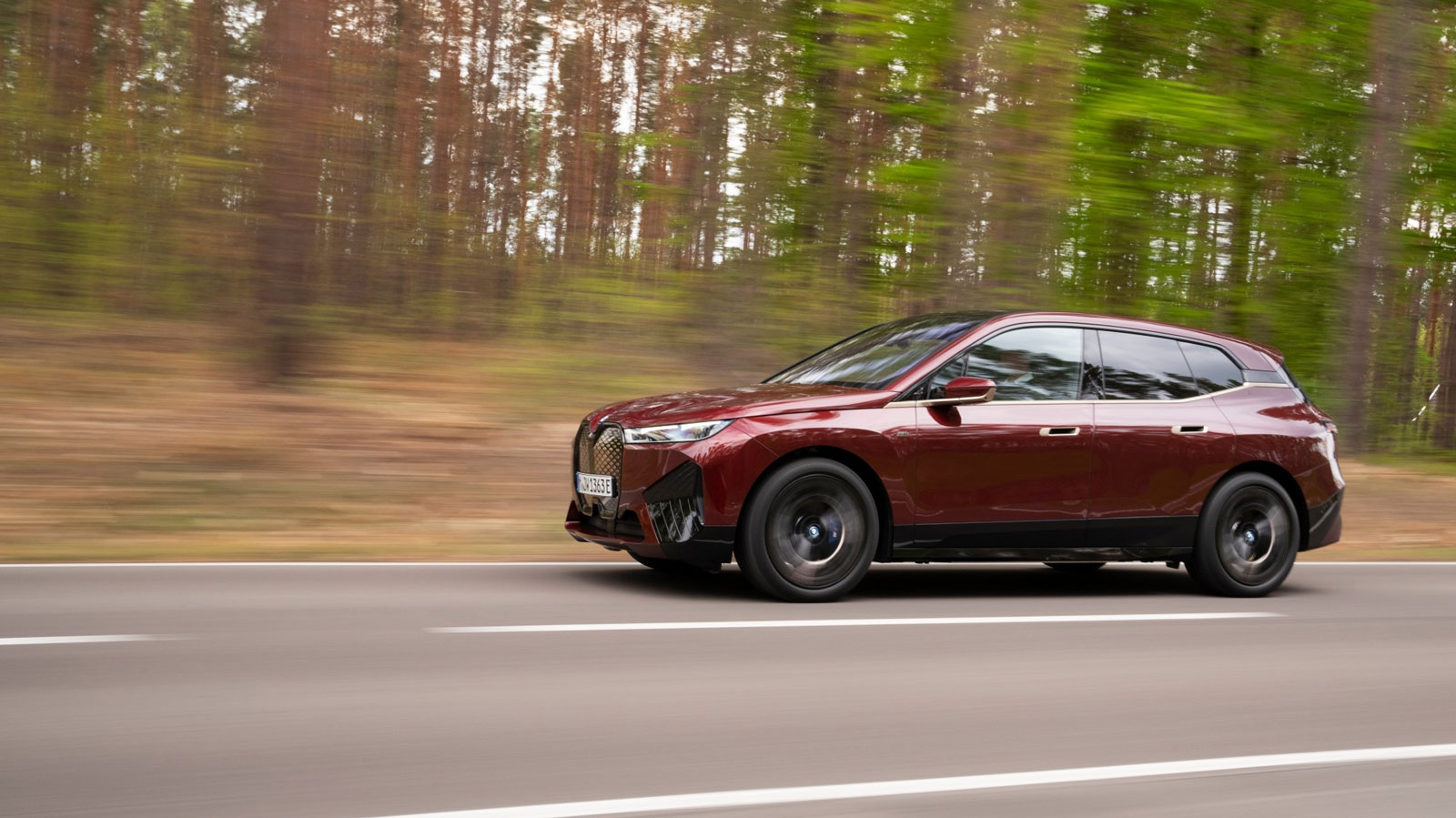 A photo of a burgundy BMW iX SUV driving on a tree-lined road. 