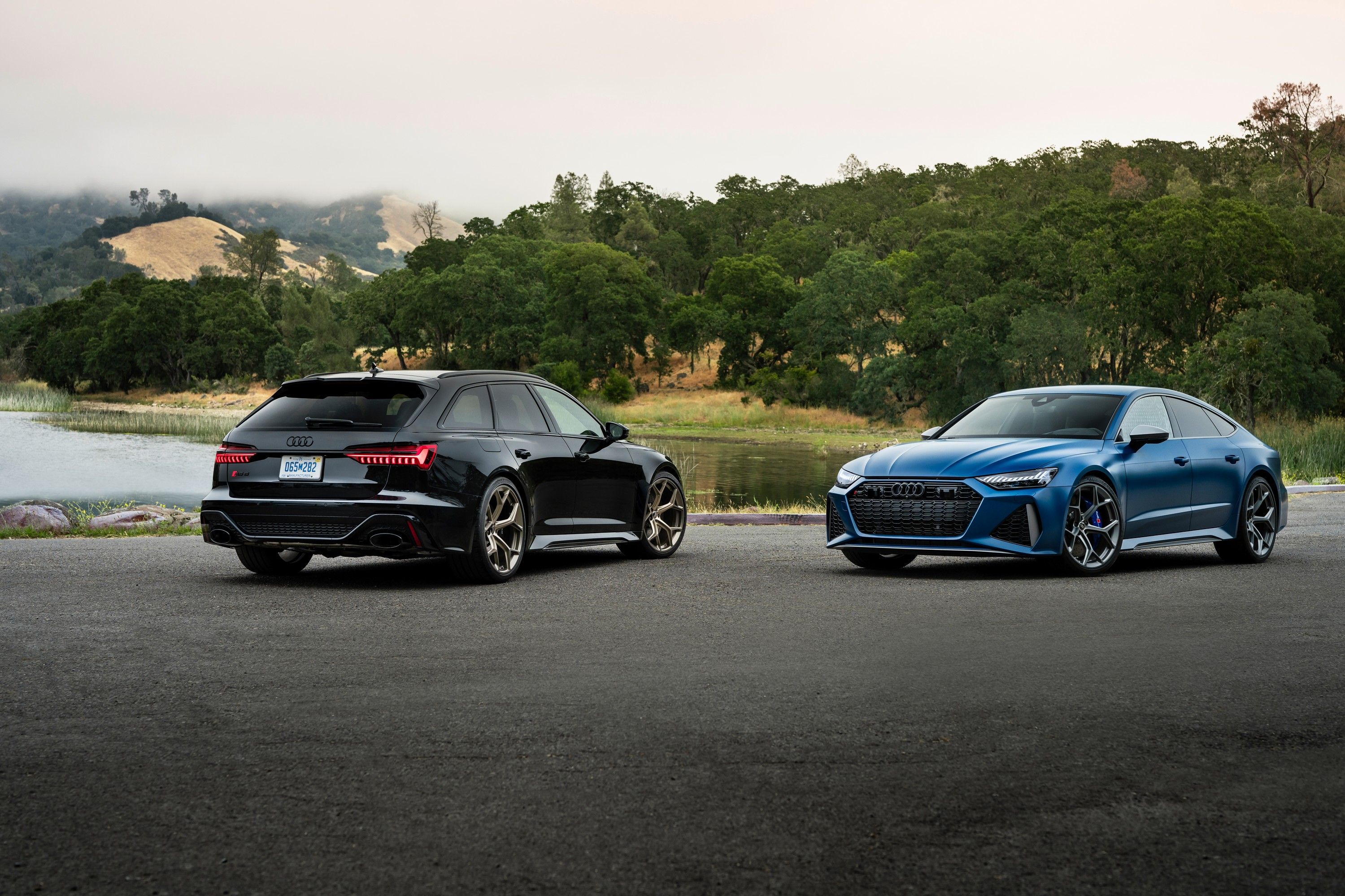 A black 2024 Audi RS 6 Avant Performance and matte blue RS 7 Performance are parked in front of a lake. The rear of the RS 6 is facing the camera, and the front three-quarter view of the RS 7 is visible.