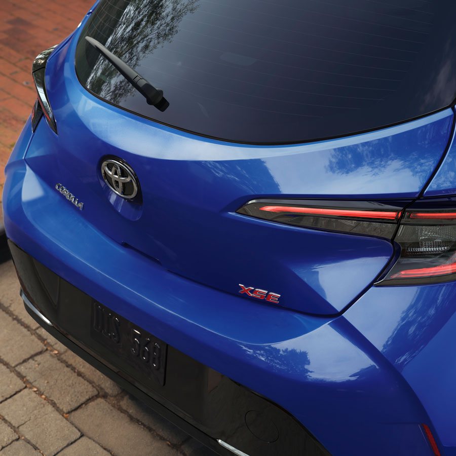 A photo of the rear end of a Toyota Corolla hatchback, the car is painted blue with a black bumper. 