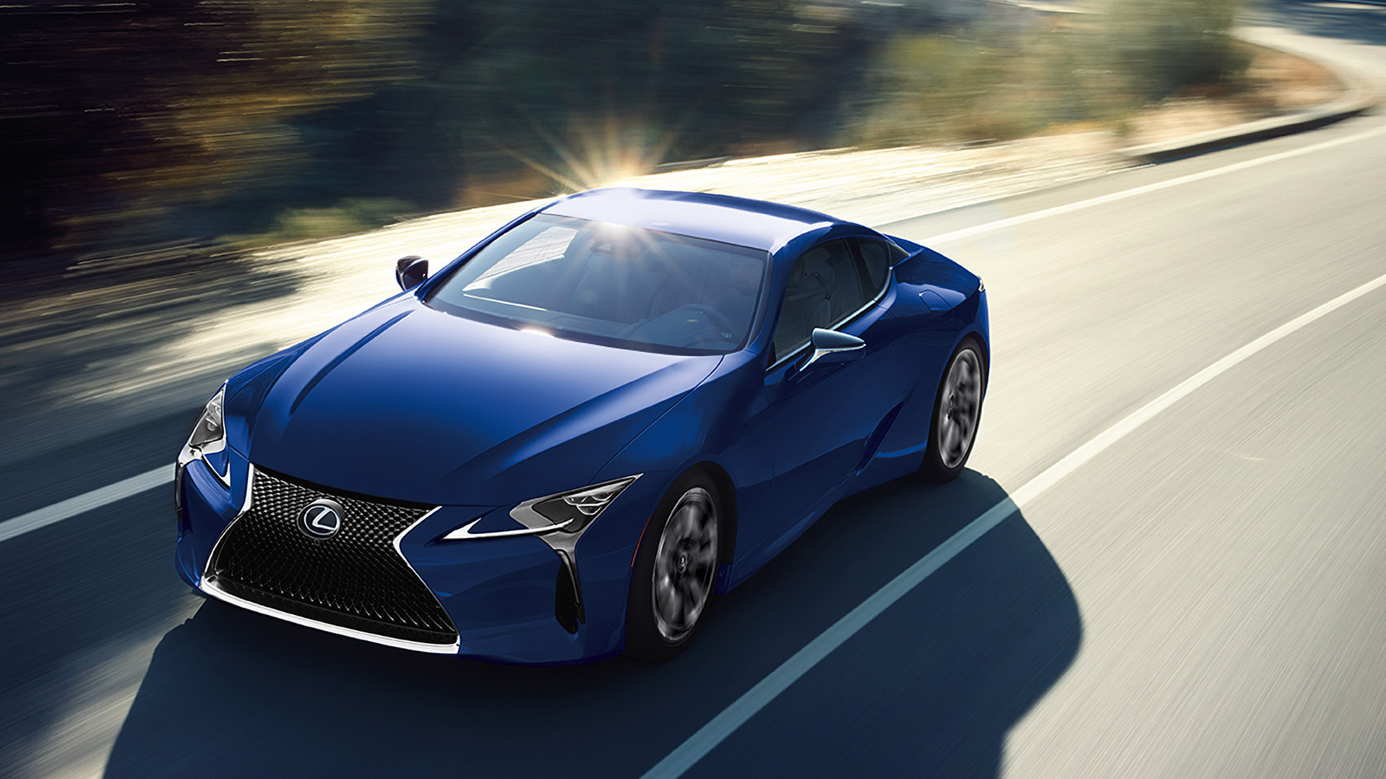 A blue 2024 Lexus LC 500h seen from the front quarter, on a road.