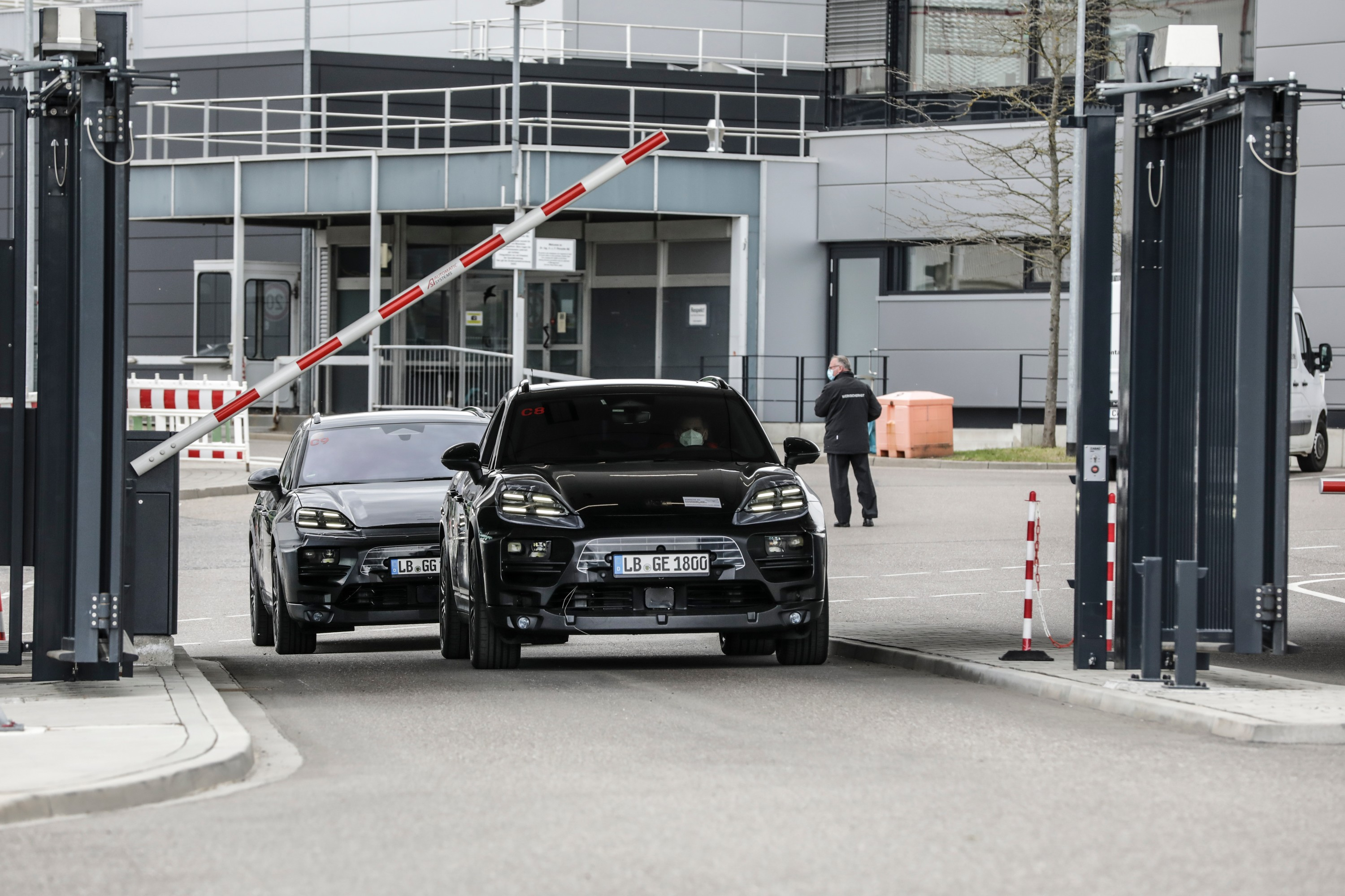 two prototypes of the porsche macan electric drive in single file through a toll booth at a test track in Germany. both vehicles are painted black and wear vinyl wrapping to disguise their design.