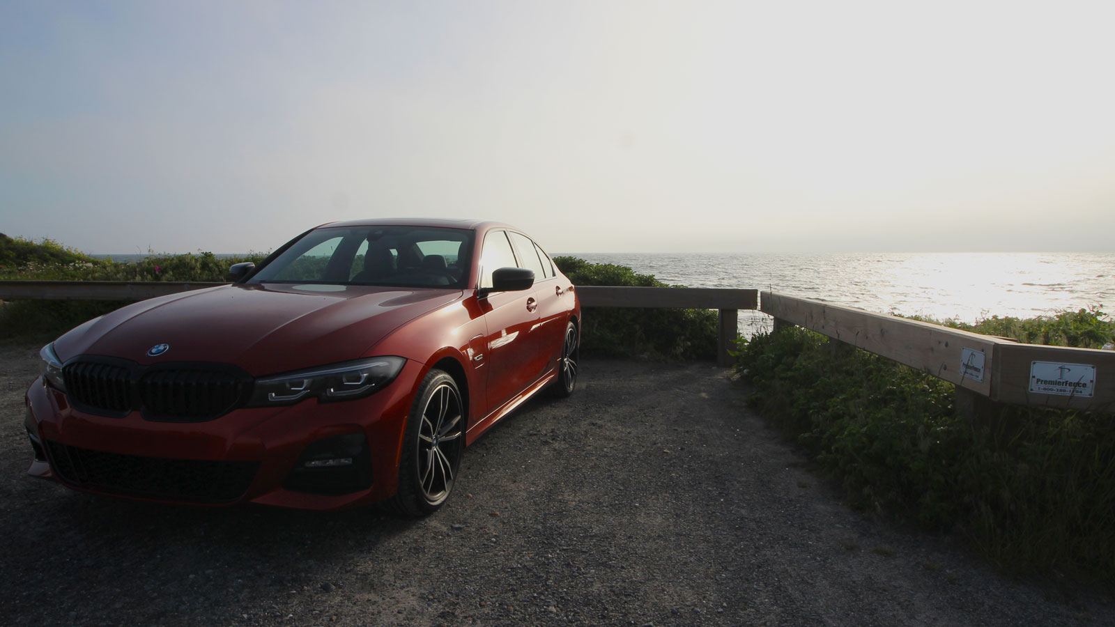 A photo of the front end of an orange BMW 330e sedan in a parking lot near the sea. 