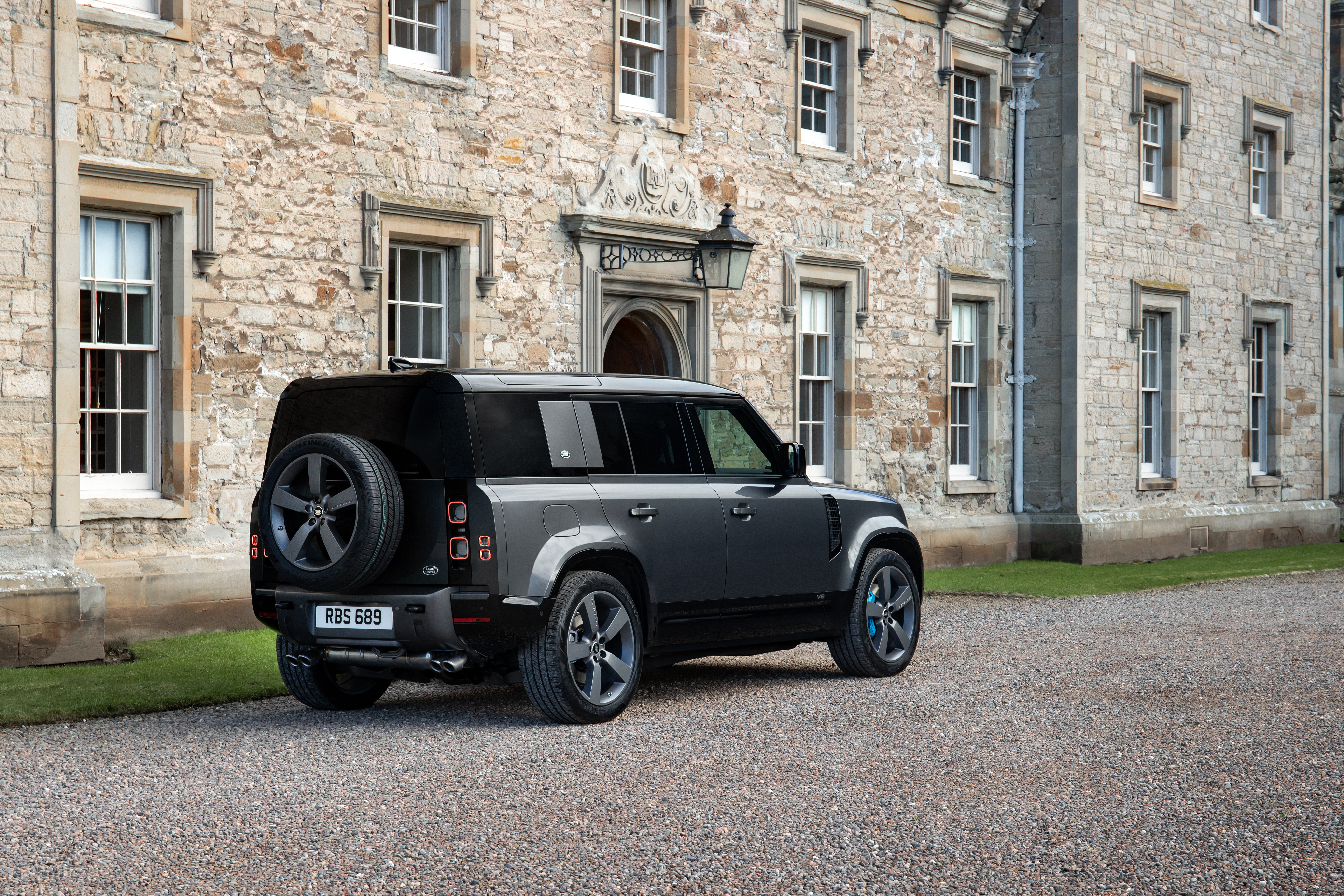 A gray land rover defender v8 is parked in front of a stately manor home