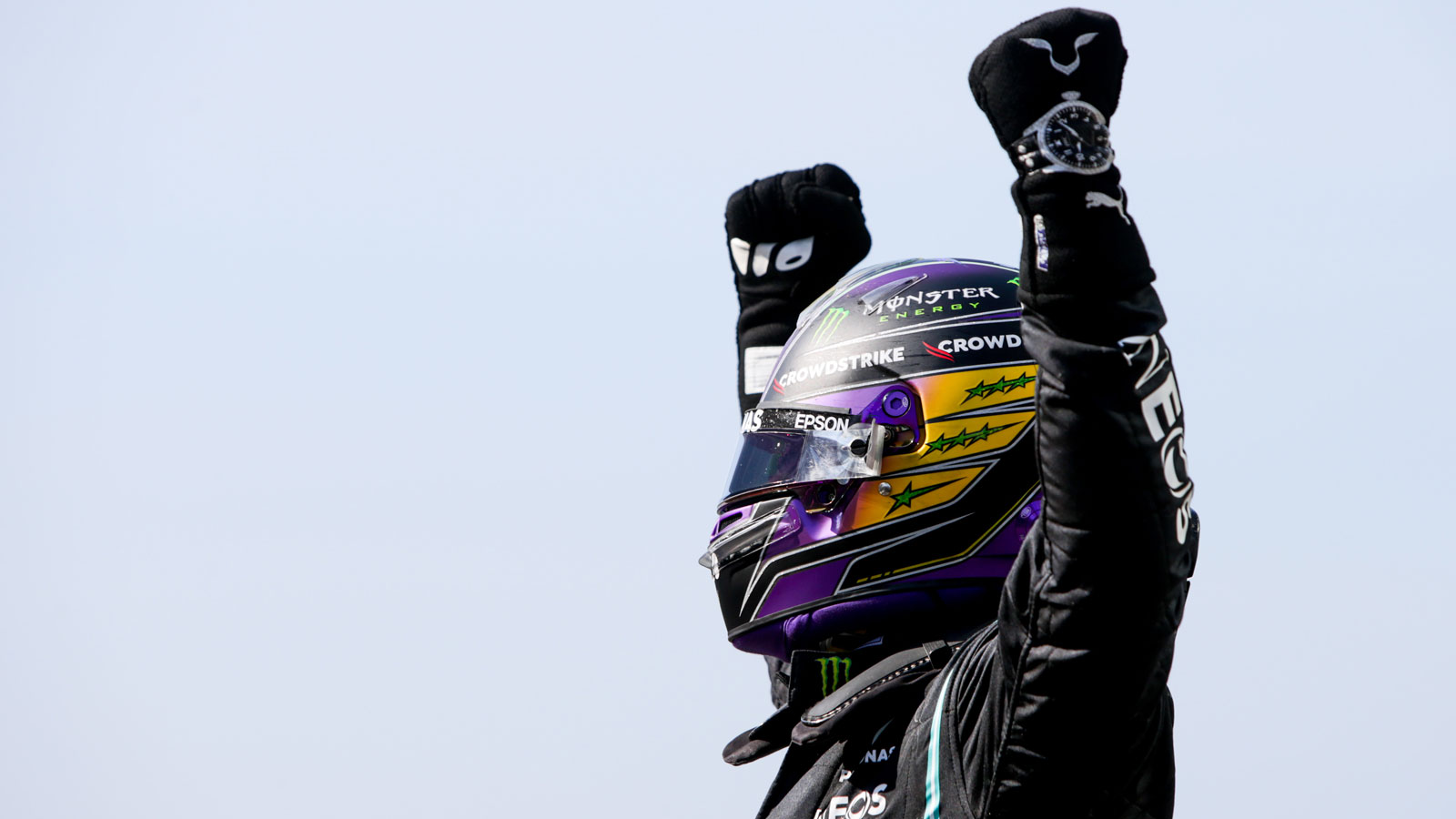 A photo of Lewis Hamilton in black racing overalls with his arms in the air. 