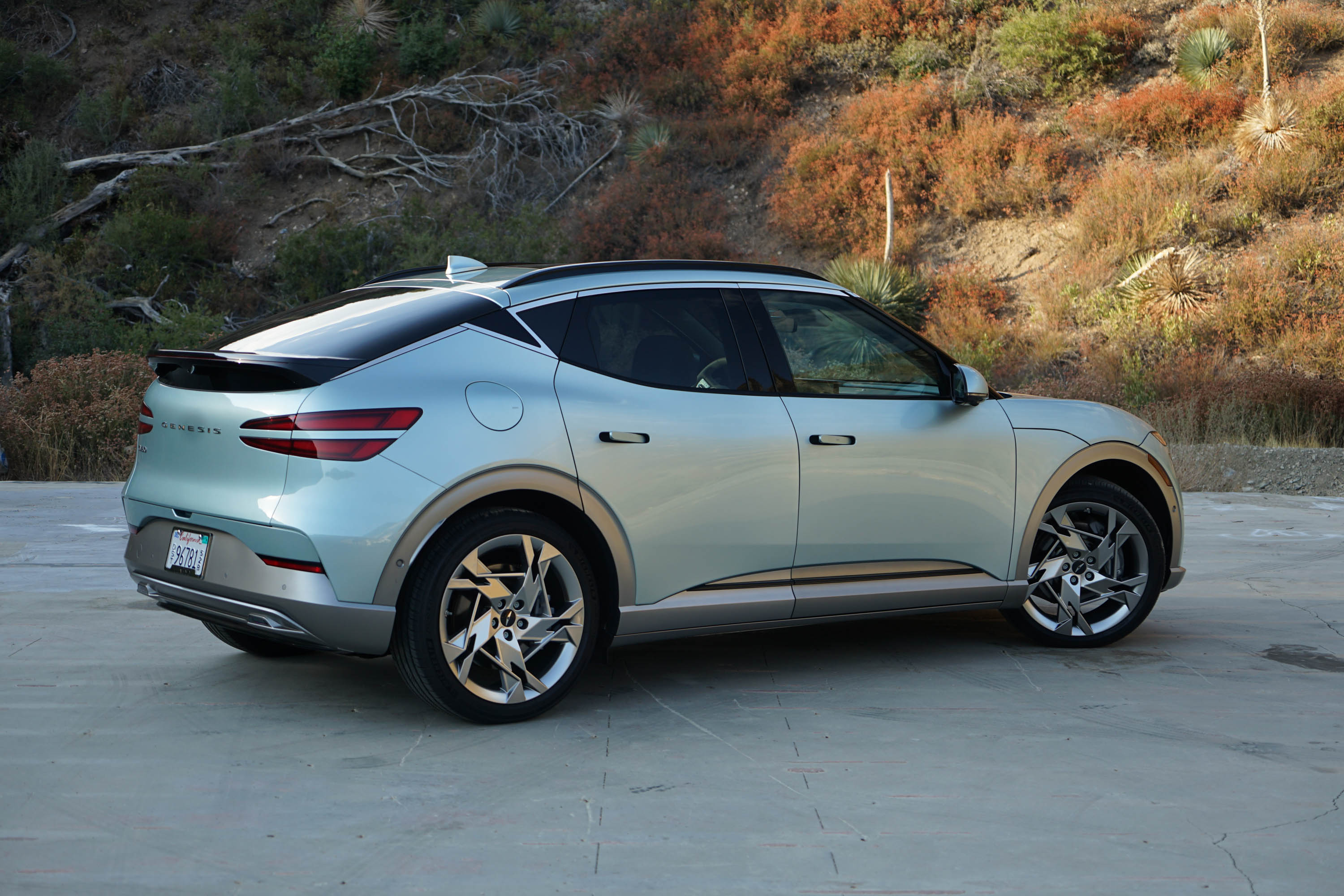 A mint green 2023 Genesis GV60 is parked in front of a hill, rear-three quarter view.