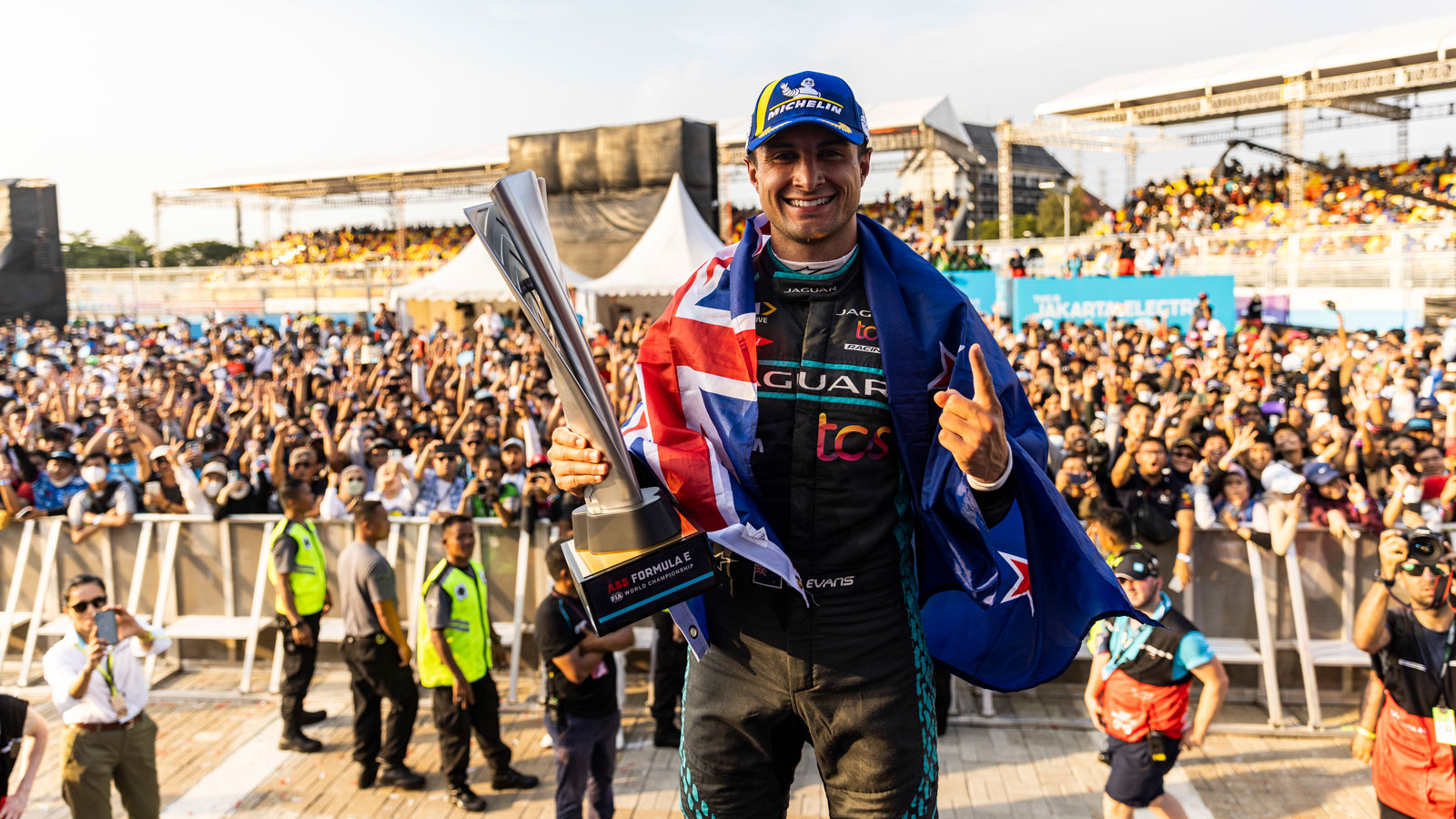 A photo of Mitch Evans on the podium in Jakarta with a New Zealand flag draped over his shoulders. 