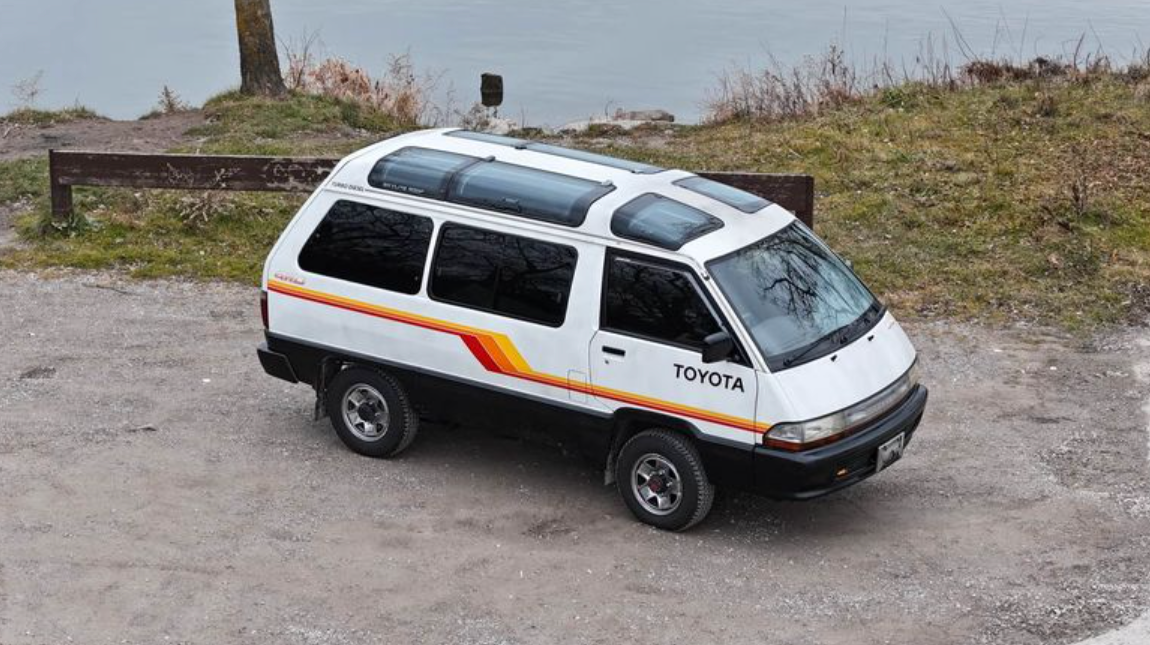 A white Toyota TownAce with orange graphics along the side sits next to a lake.