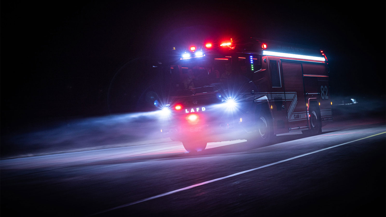 A photo of the Rosenbauer RTX fire truck driving at night with its lights on. 