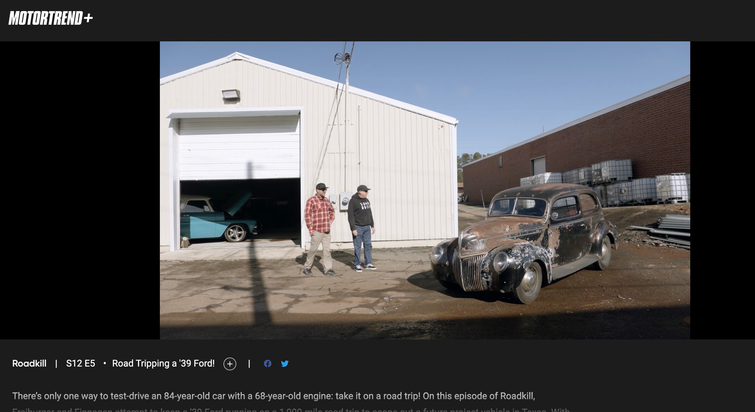 Two men stand in front of a white metal building looking at a rusty 1939 Ford Sedan