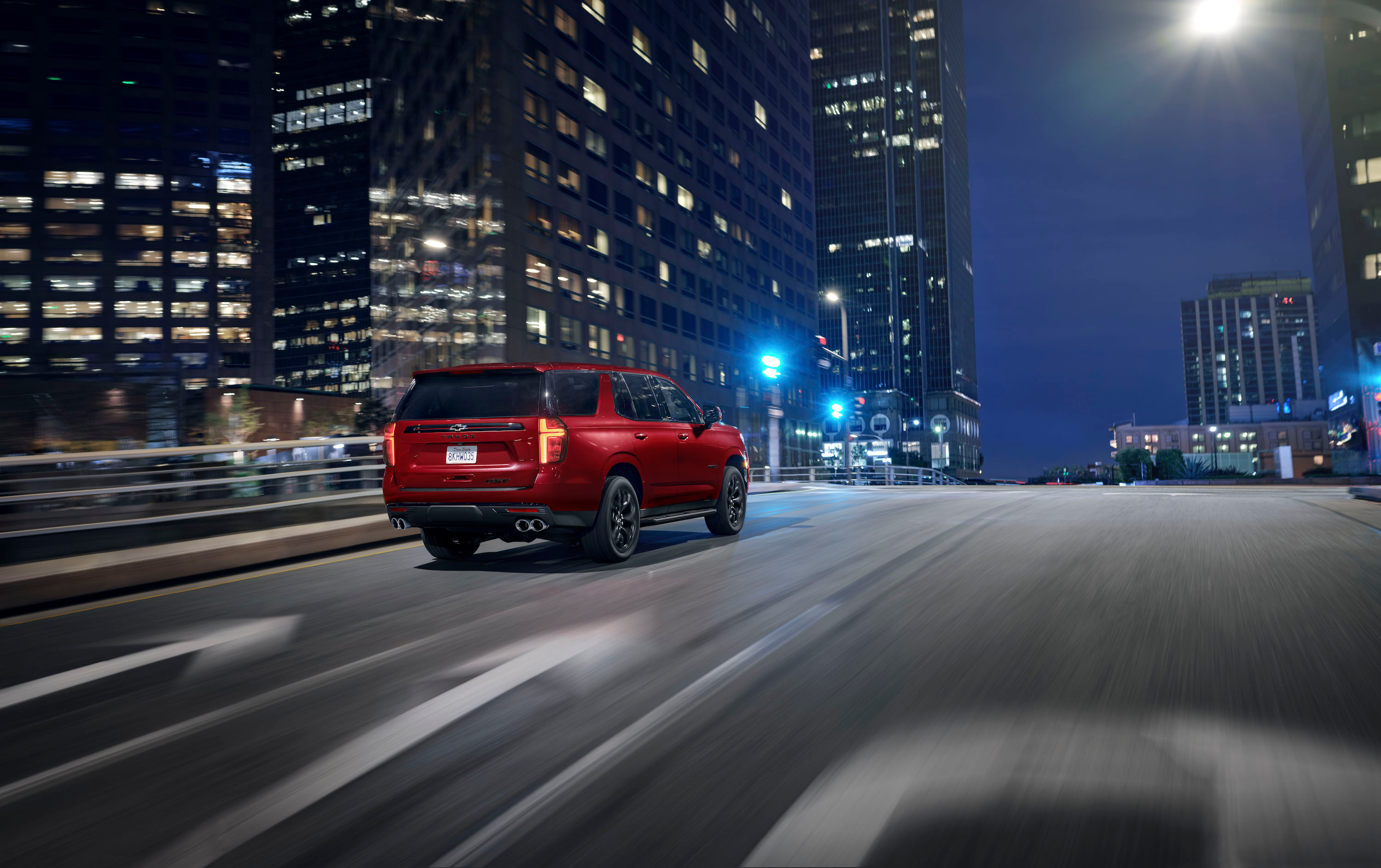 A rear view of a big red Tahoe driving on a nighttime downtown street.