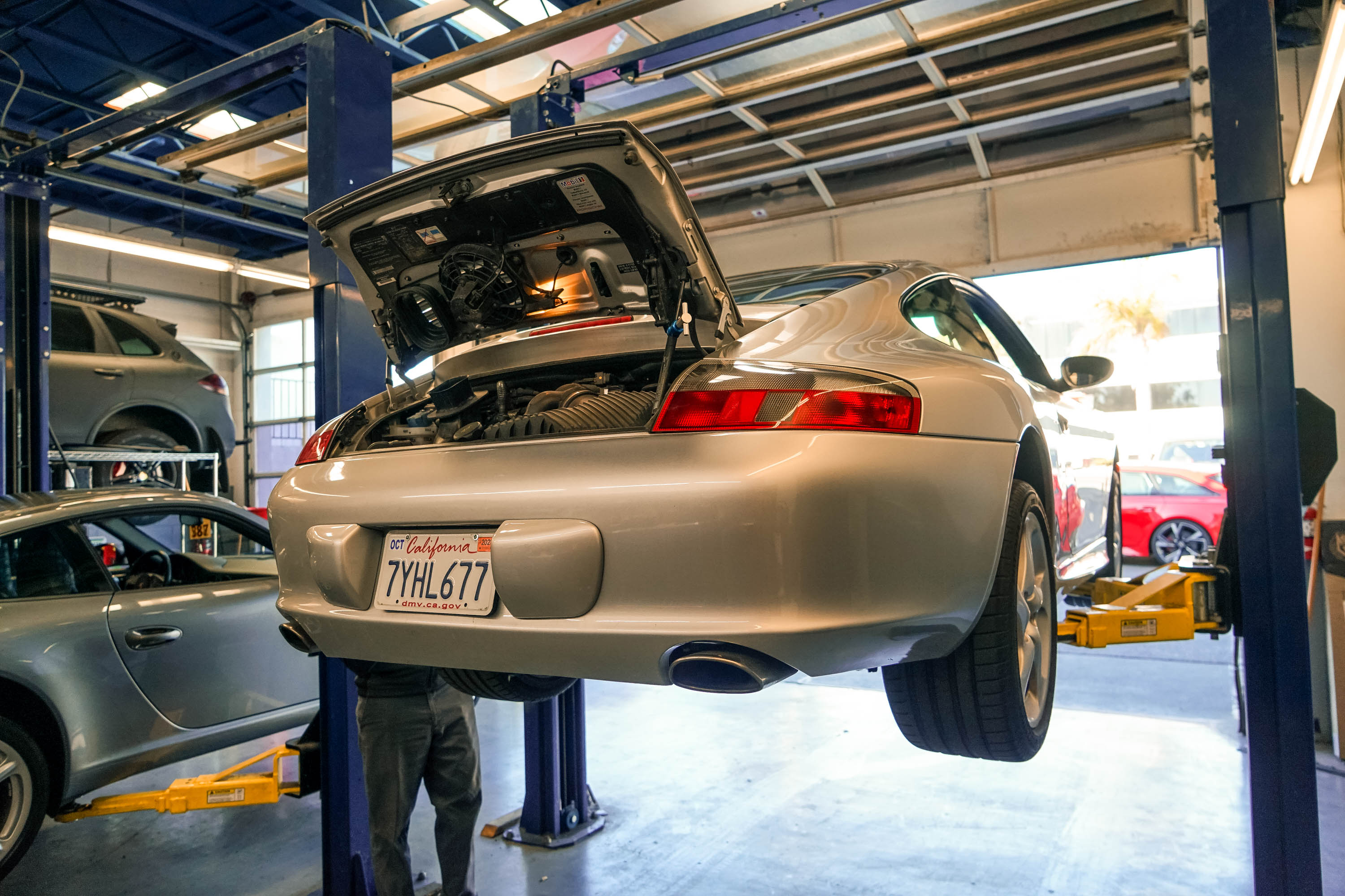 A silver 2003 Porsche 911 on a car hoist is being inspected.