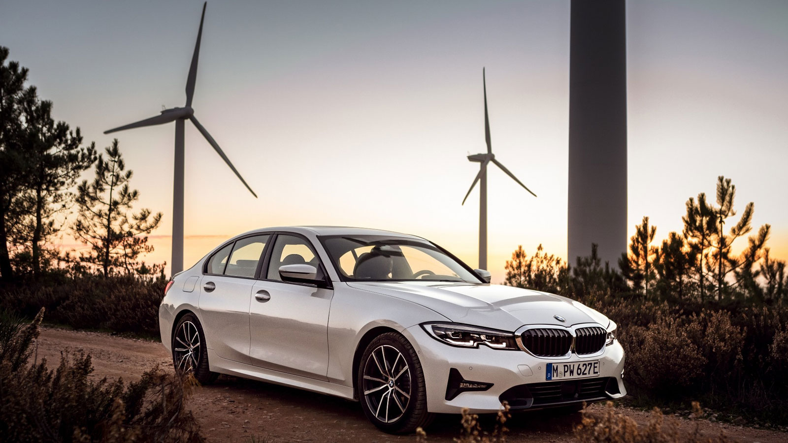 A photo of a BMW 330e sedan with wind turbines in the background. 