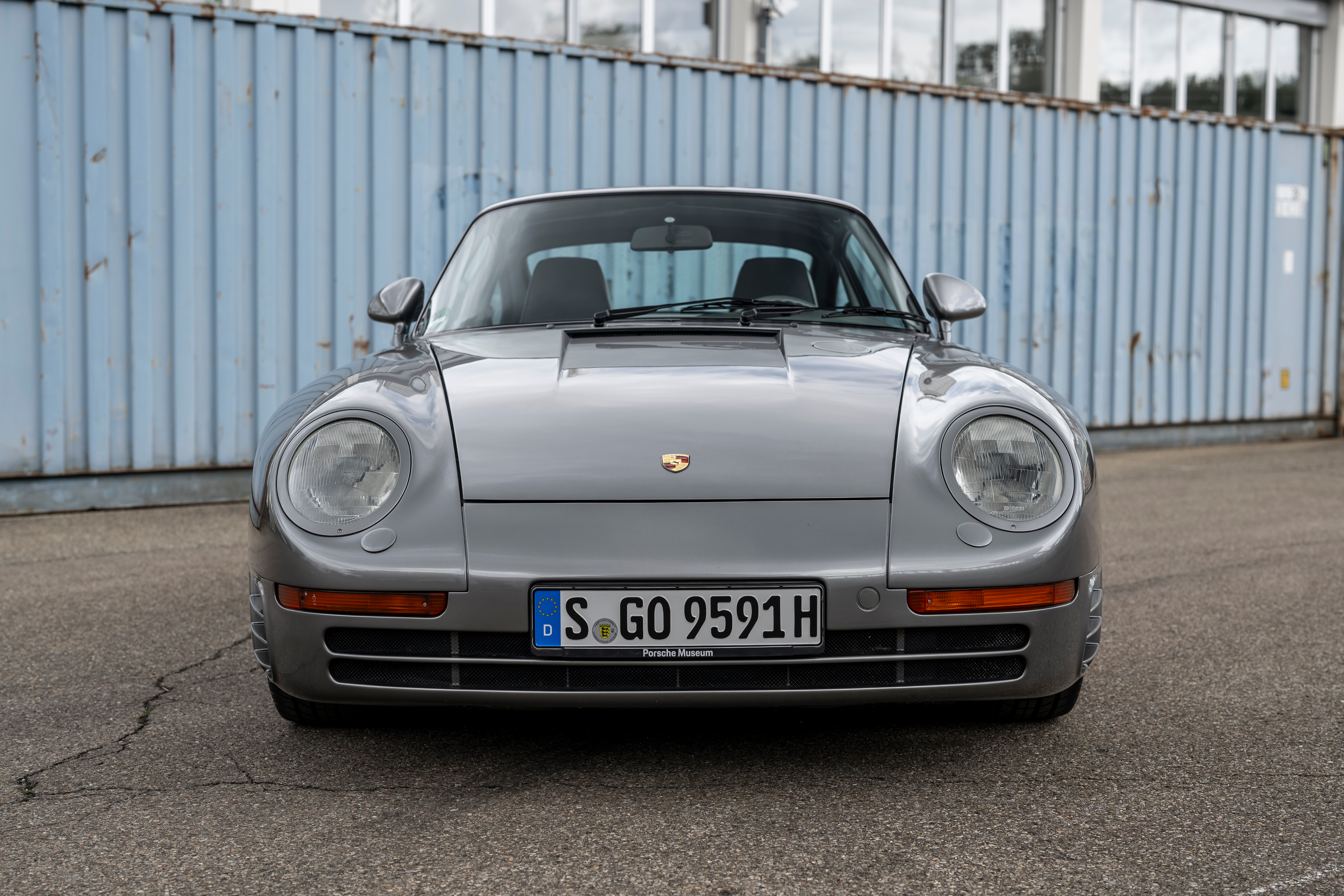 A dark silver Porsche 959 is facing the camera straight on. It's parked in front of a blue shipping container.