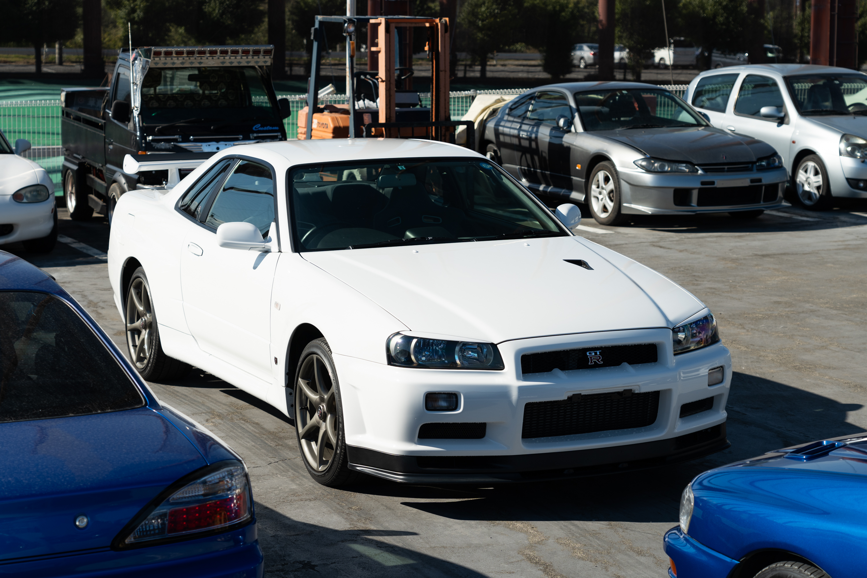 a white r34-generation nissan gt-r parked in a storage lot among numerous other JDM vehicles at an export facility in japan