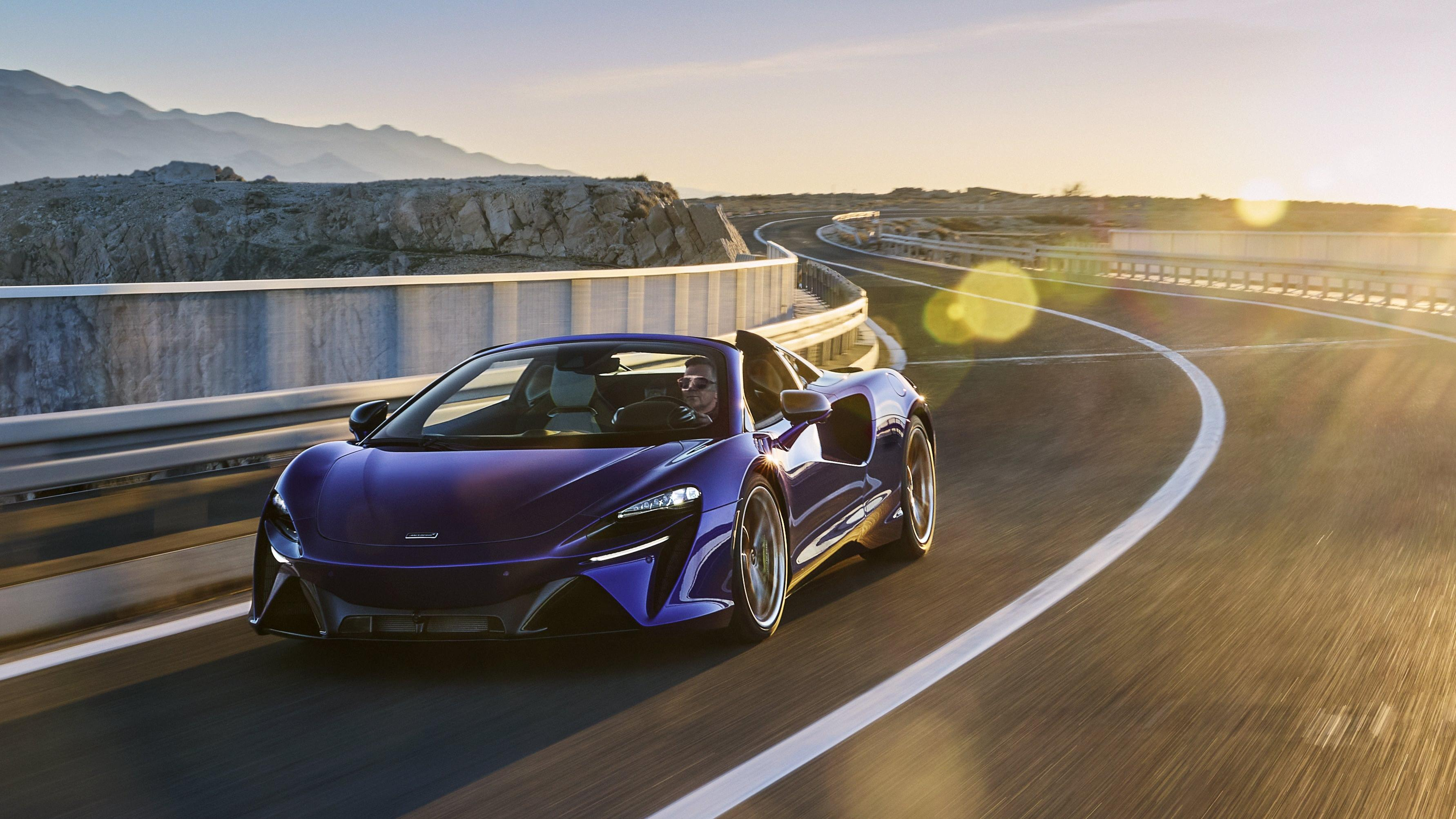 A dark blue McLaren Artura Spider driving across a winding bridge in front of mountains