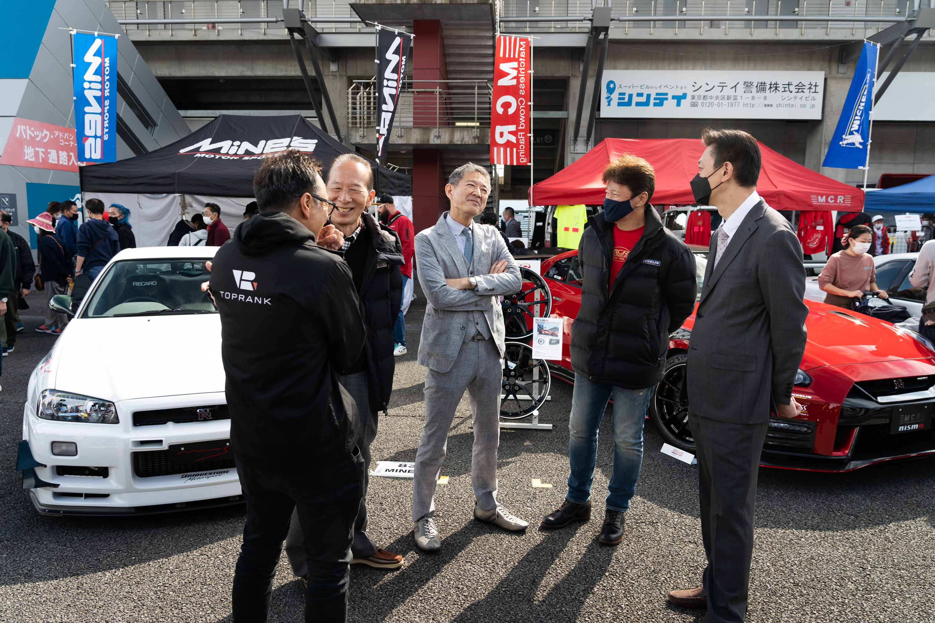 hiroshi tamura, wearing a gray tailored suit, stands in the center of a group of five men at R's Meeting, a get together for nissan gt-rs