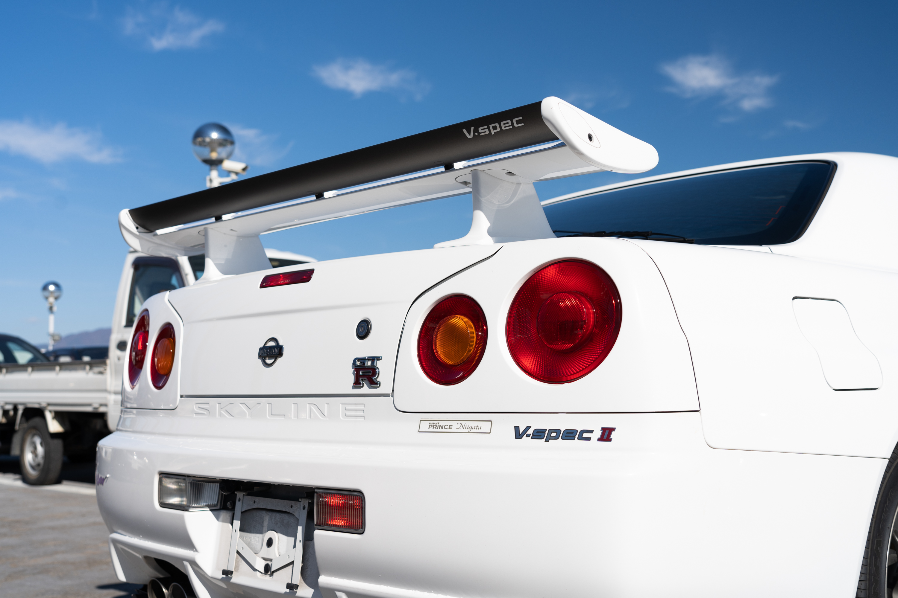 a low-angle shot of a white r34-generation nissan skyline gtr v spec ii, viewed from the right-rear corner. the car is painted white, and the large adjustable rear wing is prominently framed