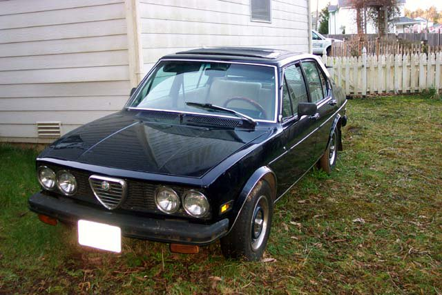 A blue 1977 Alfa Romeo Alfetta sedan is parked on grass behind a picket fence.